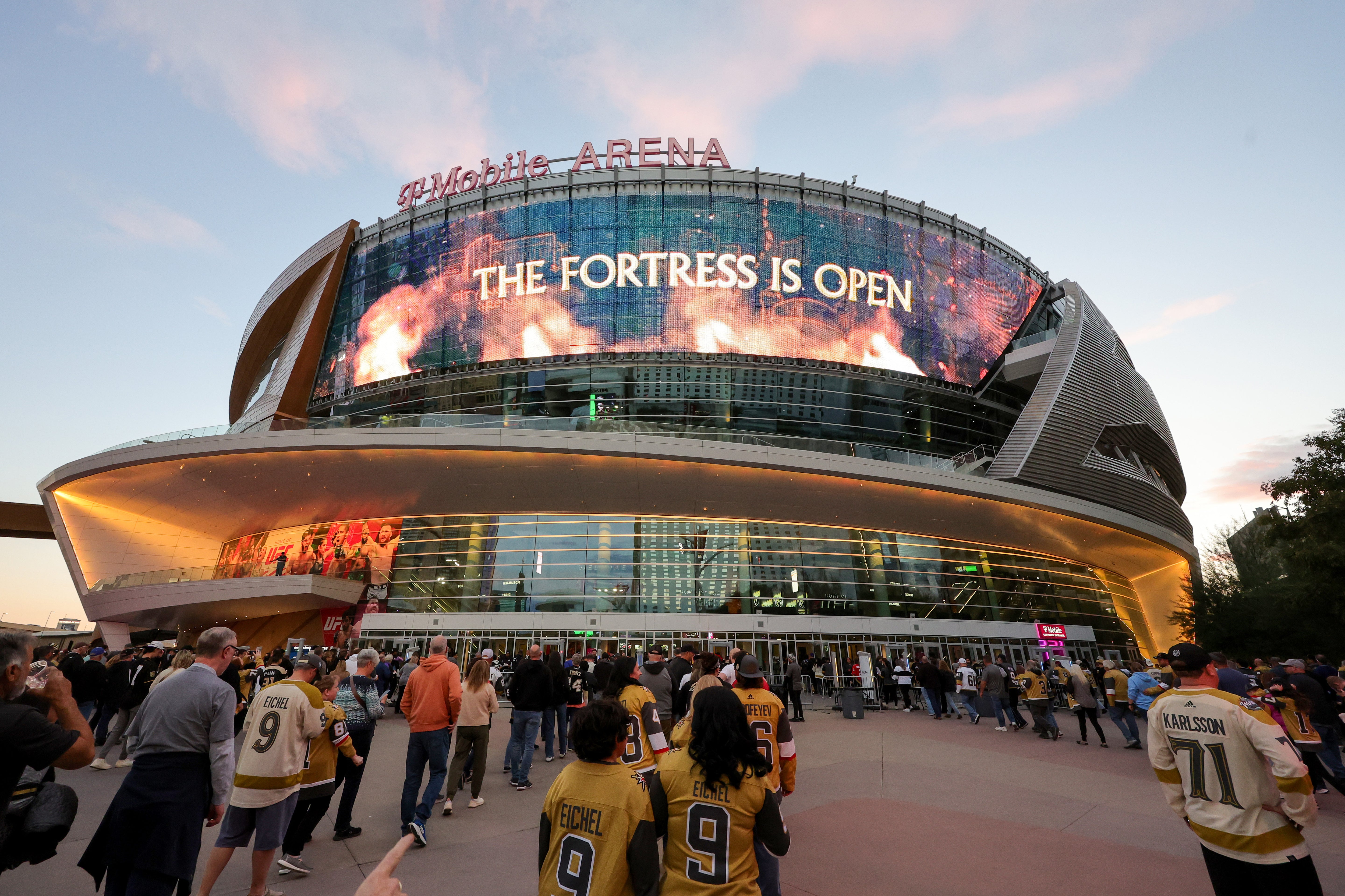 Stadium with 'The Fortress is Open' on the screen, surrounded by people in sports jerseys.